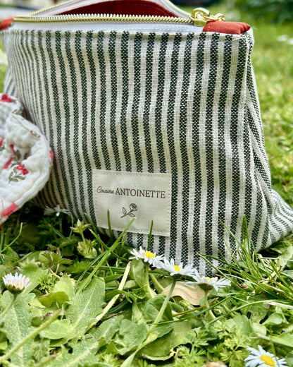 Trousse zippée Deauville Safran en coton rayé avec étiquette Cousine Antoinette, posée sur l'herbe
